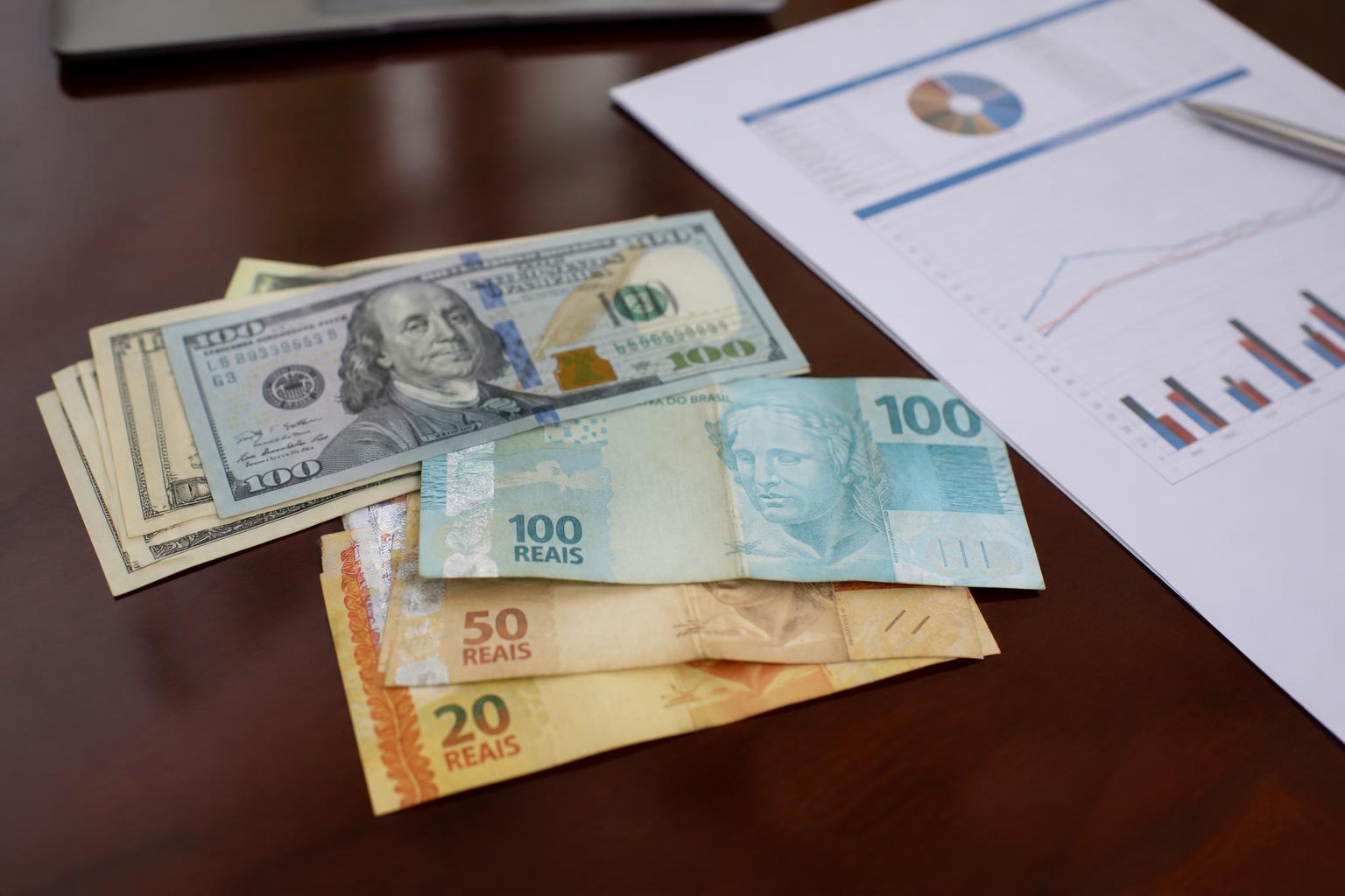 Cédulas de dólar e real sobre mesa corporativa para representar oscilação cambial